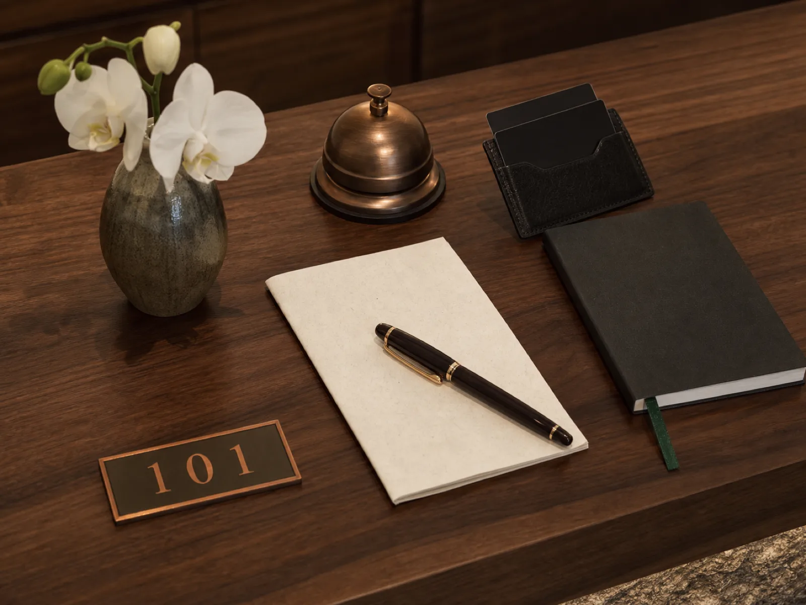 Hotel reception desk details — brass bell, key cards, guest folio, fountain pen, fresh orchid — arranged on polished walnut